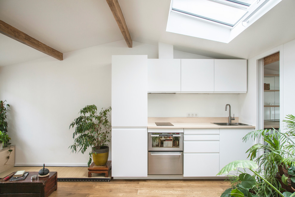 Example of a small danish single-wall light wood floor open concept kitchen design in Paris with a drop-in sink, flat-panel cabinets, white cabinets, stainless steel appliances, no island and beige countertops