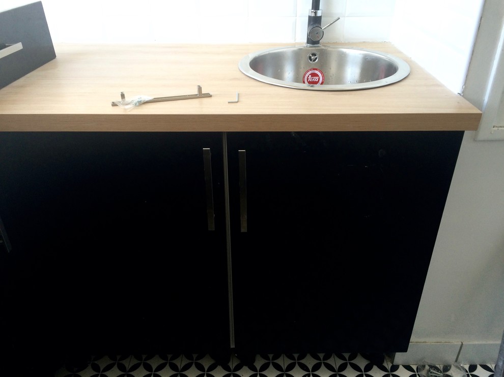 Bild på ett avskilt, litet funkis l-kök, med en undermonterad diskho, släta luckor, svarta skåp, träbänkskiva, vitt stänkskydd, stänkskydd i tunnelbanekakel, integrerade vitvaror och linoleumgolv