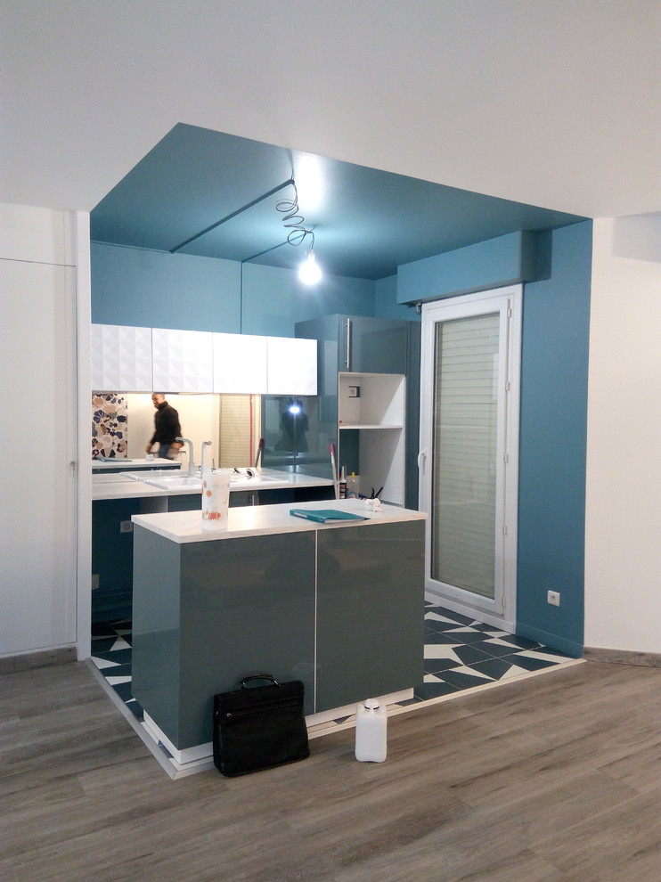 Modern inredning av ett mellanstort vit vitt kök, med en nedsänkt diskho, släta luckor, blå skåp, marmorbänkskiva, stänkskydd med metallisk yta, spegel som stänkskydd, vita vitvaror, mörkt trägolv, en köksö och blått golv