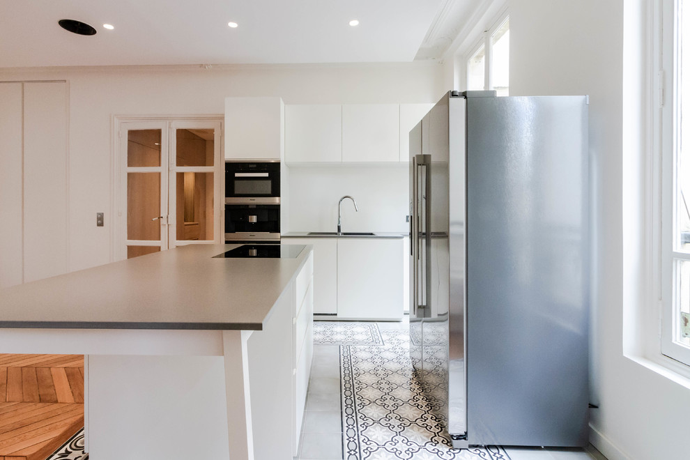 Foto på ett mellanstort funkis grå linjärt kök med öppen planlösning, med en integrerad diskho, släta luckor, vita skåp, granitbänkskiva, vitt stänkskydd, glaspanel som stänkskydd, rostfria vitvaror, cementgolv, en köksö och grått golv