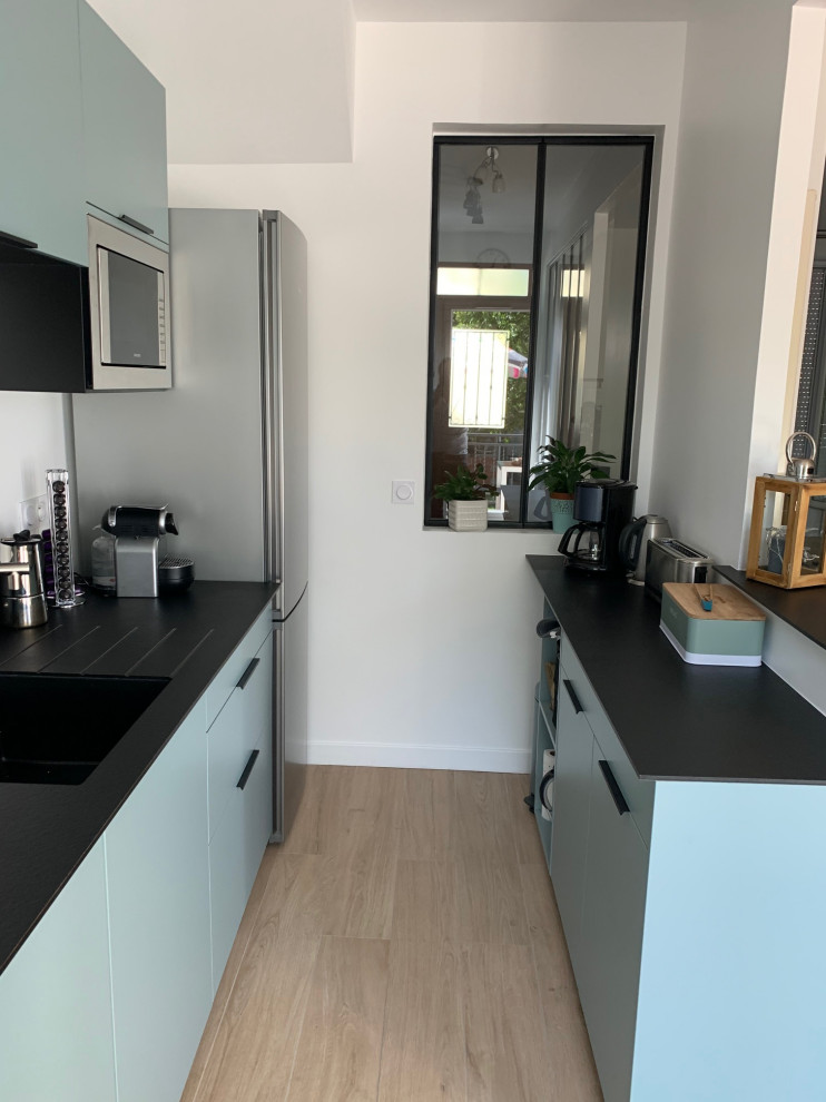 Photo of a medium sized modern galley open plan kitchen with a submerged sink, flat-panel cabinets, green cabinets, granite worktops, black splashback, granite splashback, stainless steel appliances, light hardwood flooring, an island, brown floors, black worktops and a wallpapered ceiling.