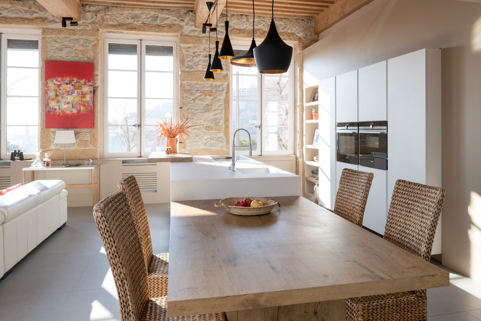 На фото: кухня-гостиная в средиземноморском стиле с одинарной мойкой, плоскими фасадами, белыми фасадами, черной техникой, островом, серым полом, белой столешницей и окном