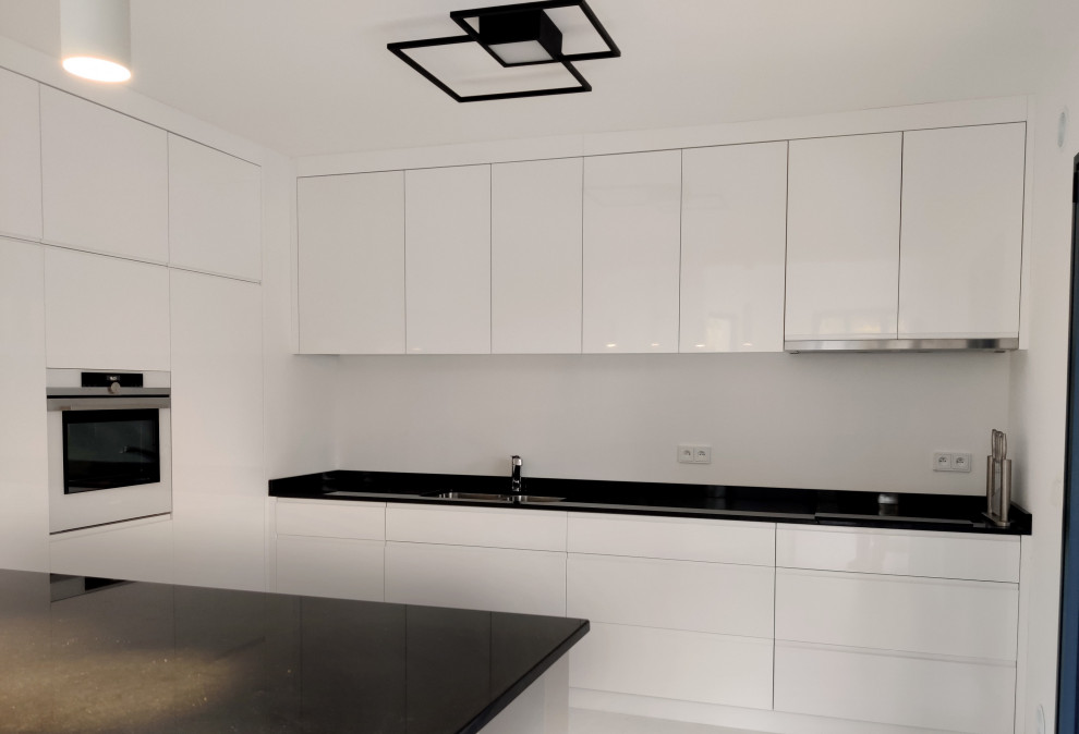 Photo of a large contemporary l-shaped kitchen/diner in Other with an integrated sink, beaded cabinets, white cabinets, granite worktops, white splashback, integrated appliances, concrete flooring, an island, white floors and black worktops.