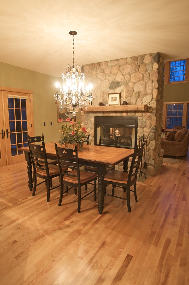 Cette photo montre une salle à manger ouverte sur la cuisine montagne de taille moyenne avec un mur vert, parquet en bambou, une cheminée double-face et un manteau de cheminée en pierre.