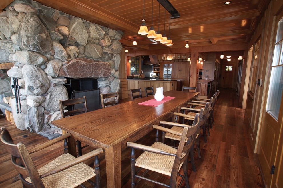 Immagine di una grande sala da pranzo aperta verso la cucina rustica con pareti marroni, pavimento in legno massello medio, camino sospeso e cornice del camino in pietra