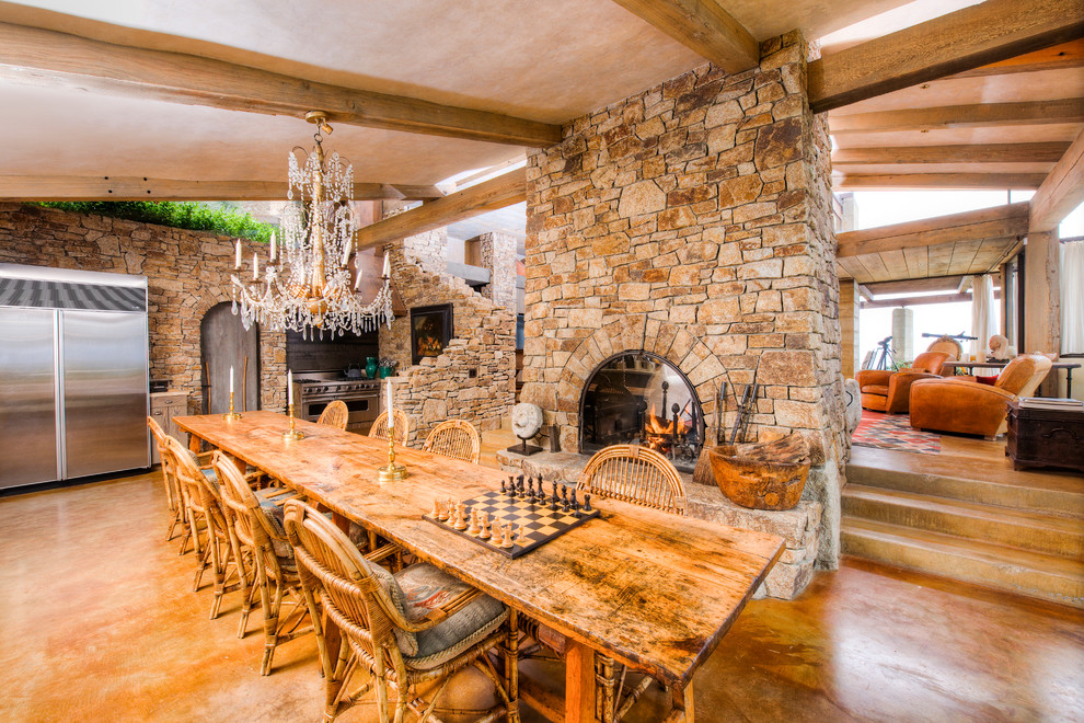 На фото: огромная кухня-столовая в средиземноморском стиле с коричневыми стенами, бетонным полом, двусторонним камином и фасадом камина из камня с