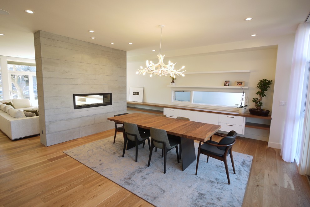 Cette image montre une salle à manger ouverte sur la cuisine minimaliste de taille moyenne avec un mur blanc, un sol en bois brun, une cheminée double-face et un manteau de cheminée en béton.