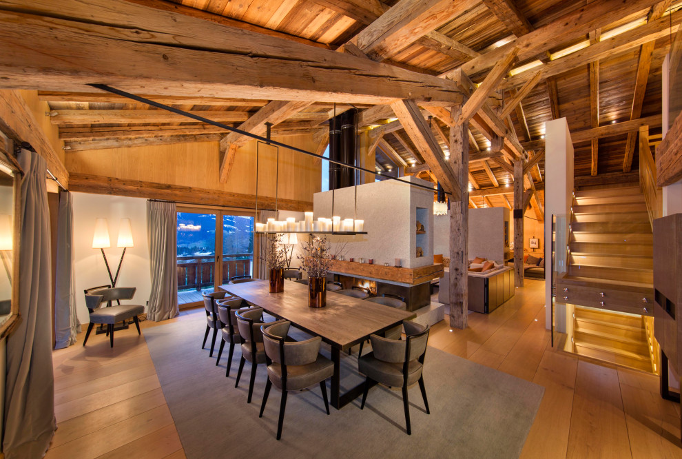 Cette photo montre une salle à manger ouverte sur le salon montagne avec une cheminée double-face, poutres apparentes, un plafond voûté, un plafond en bois, un mur blanc, un sol en bois brun et un sol marron.