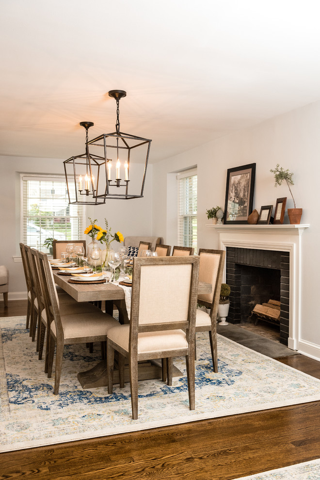Cette photo montre une salle à manger ouverte sur le salon chic avec un mur blanc, un sol en bois brun, une cheminée standard, un manteau de cheminée en brique et un sol marron.