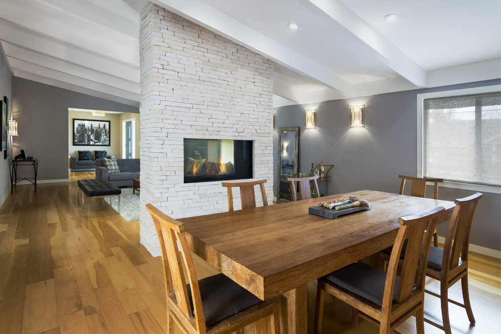 Esempio di una grande sala da pranzo aperta verso la cucina minimalista con parquet chiaro, camino bifacciale, cornice del camino in pietra e pareti grigie