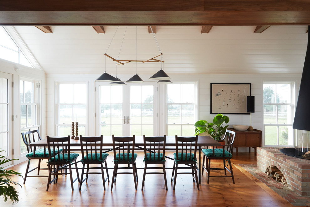 Cette photo montre une salle à manger bord de mer avec un mur blanc, un sol en bois brun et cheminée suspendue.