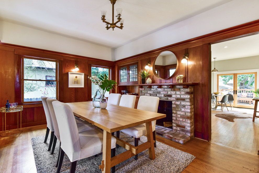 Cette photo montre une salle à manger craftsman en bois fermée avec un mur blanc, un sol en bois brun, une cheminée standard, un manteau de cheminée en brique et un sol marron.