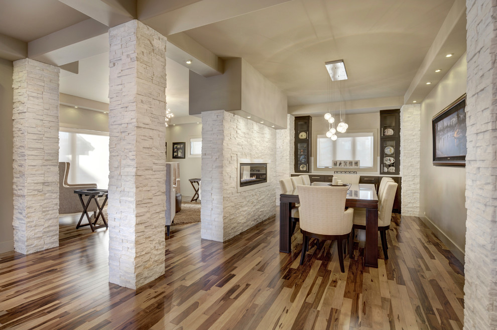 Cette image montre une salle à manger ouverte sur la cuisine minimaliste de taille moyenne avec un mur beige, un sol en bois brun, une cheminée double-face et un manteau de cheminée en pierre.