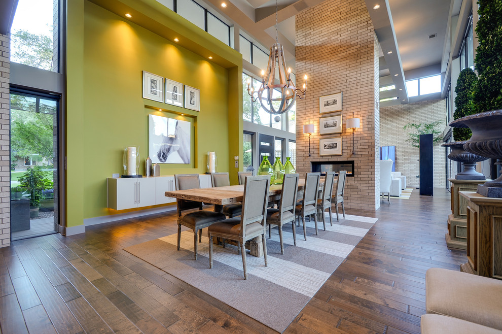 Cette photo montre une grande salle à manger ouverte sur le salon moderne avec un mur vert, un sol en bois brun, une cheminée double-face et un manteau de cheminée en brique.