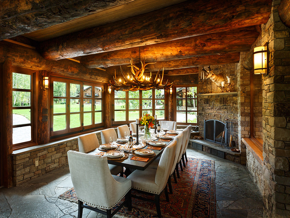 Cette photo montre une grande salle à manger ouverte sur le salon montagne avec un mur gris, un sol en ardoise, une cheminée d'angle et un manteau de cheminée en pierre.