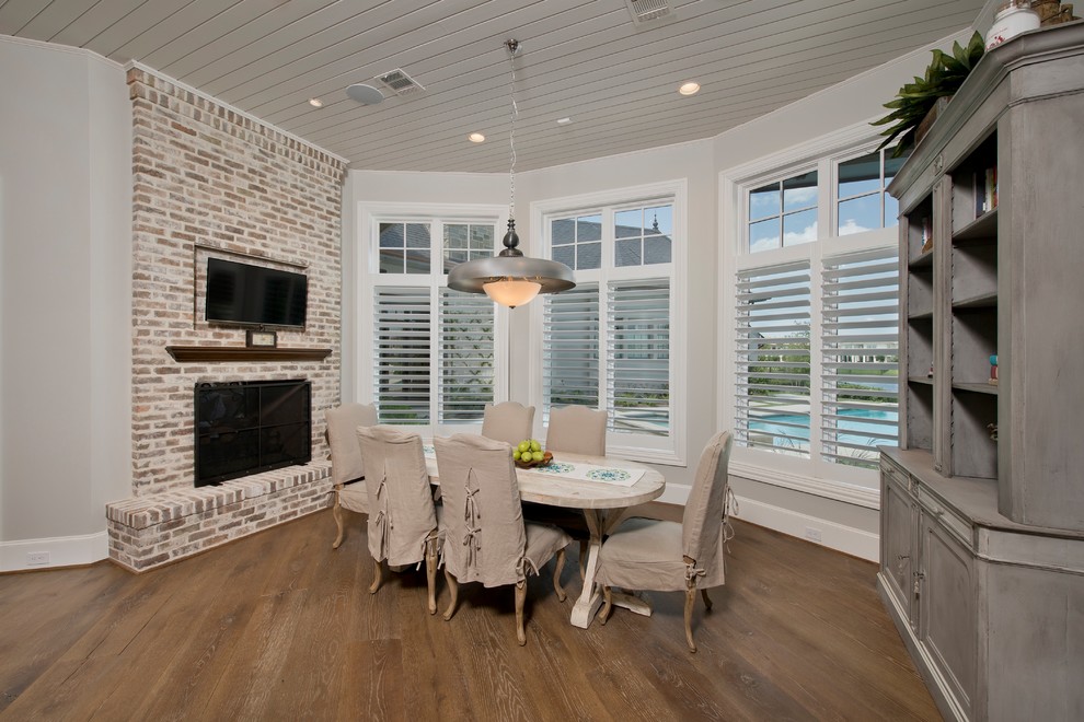Mittelgroßes Landhaus Esszimmer mit grauer Wandfarbe, braunem Holzboden, Kamin, Kaminumrandung aus Backstein und braunem Boden in Houston