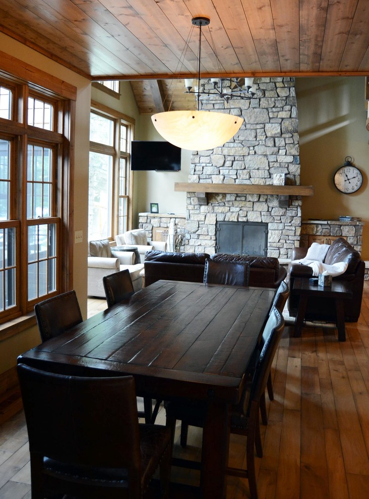 Cette image montre une grande salle à manger ouverte sur le salon chalet avec un mur beige, un sol en bois brun, une cheminée standard et un manteau de cheminée en pierre.