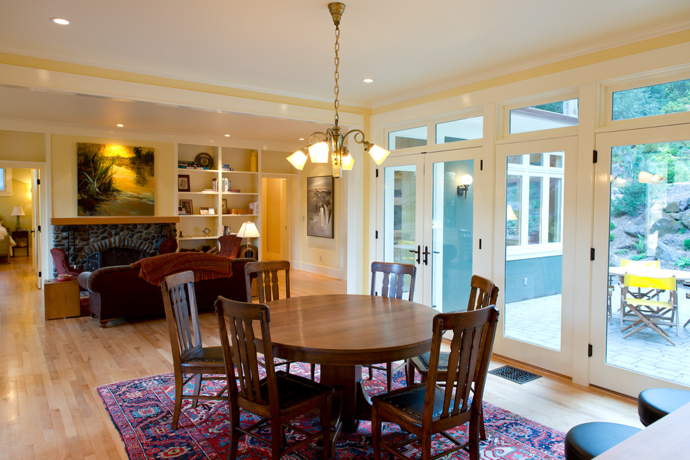 Cette photo montre une salle à manger ouverte sur le salon craftsman de taille moyenne avec un mur jaune, parquet clair, une cheminée standard et un manteau de cheminée en pierre.