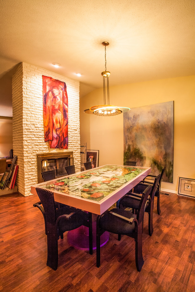 Cette photo montre une salle à manger ouverte sur le salon tendance de taille moyenne avec un mur jaune, sol en stratifié, une cheminée double-face et un manteau de cheminée en brique.