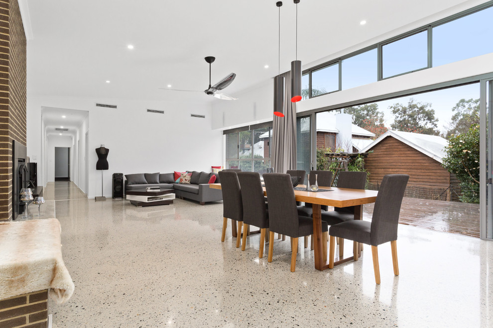 Immagine di una grande sala da pranzo aperta verso il soggiorno moderna con pareti bianche, pavimento in cemento, camino classico, cornice del camino in mattoni e pavimento grigio
