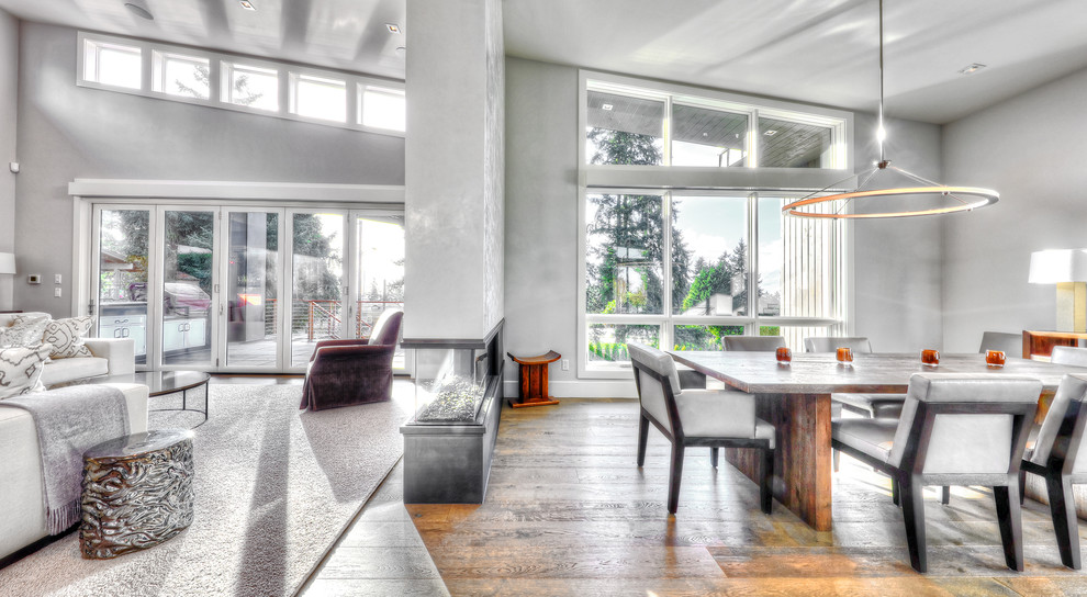 Aménagement d'une grande salle à manger ouverte sur le salon contemporaine avec un mur gris, un sol en bois brun, une cheminée double-face, un manteau de cheminée en métal et éclairage.