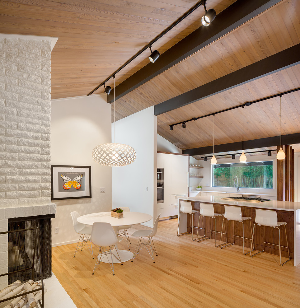 Immagine di una sala da pranzo aperta verso la cucina moderna con pareti bianche, pavimento in legno massello medio, camino bifacciale, cornice del camino in mattoni e pavimento bianco