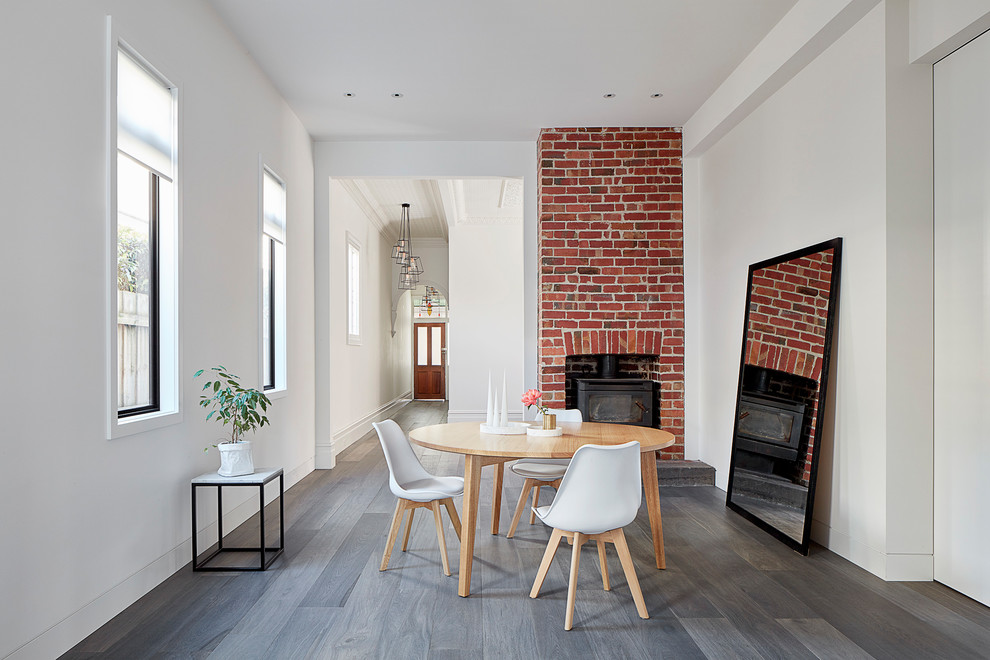 Idée de décoration pour une salle à manger ouverte sur la cuisine design de taille moyenne avec un mur blanc, parquet clair, un poêle à bois, un manteau de cheminée en brique et un sol gris.