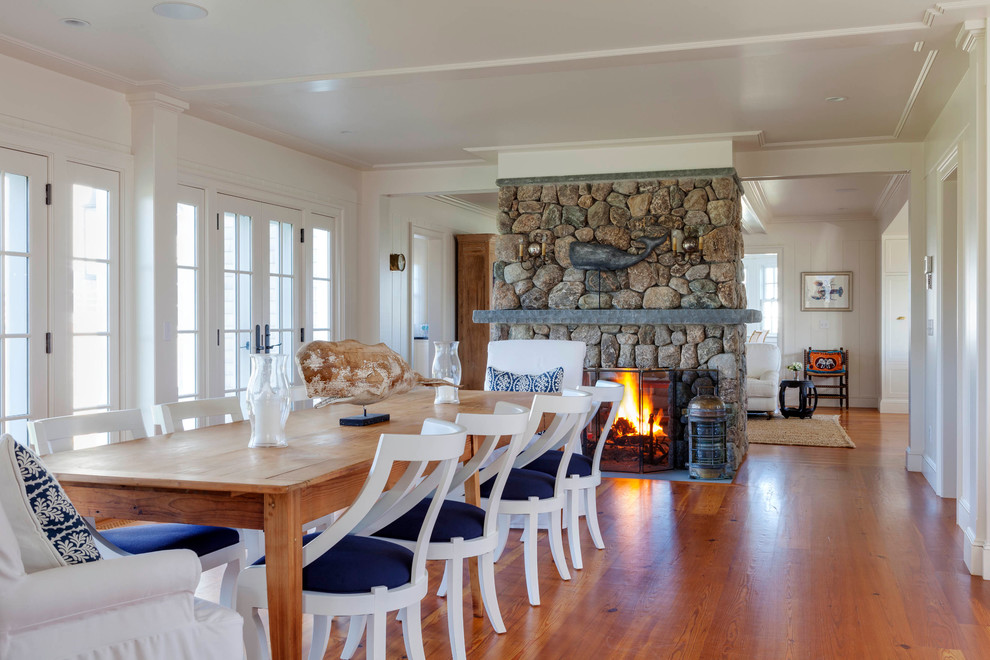 Cette photo montre une grande salle à manger ouverte sur le salon bord de mer avec un mur blanc, une cheminée double-face, un manteau de cheminée en pierre et parquet clair.
