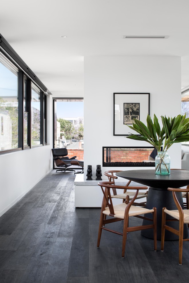 Cette photo montre une salle à manger ouverte sur le salon moderne de taille moyenne avec un mur blanc, parquet foncé, une cheminée double-face, un manteau de cheminée en plâtre et un sol noir.