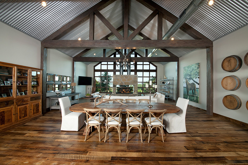 Photo of an expansive rustic open plan dining room in Austin with white walls, dark hardwood flooring, a two-sided fireplace, a stone fireplace surround, brown floors and feature lighting.