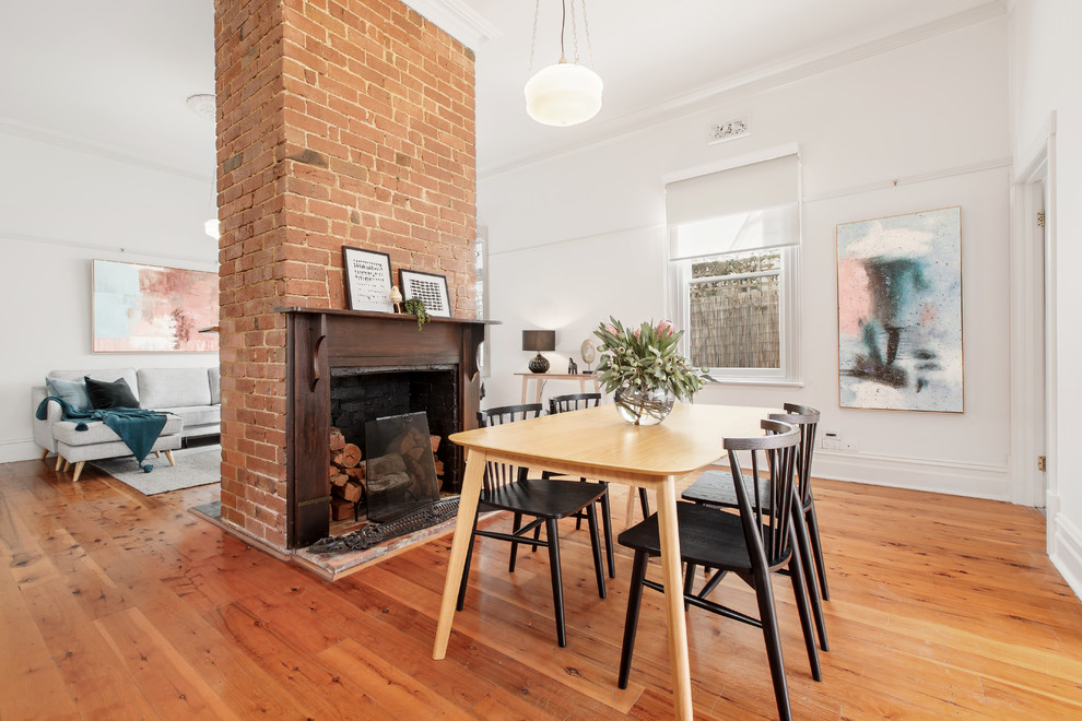 Cette photo montre une salle à manger ouverte sur le salon tendance avec un mur blanc, un sol en bois brun, une cheminée standard, un manteau de cheminée en brique et un sol marron.