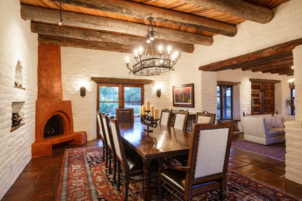 Cette image montre une grande salle à manger ouverte sur le salon sud-ouest américain avec un mur blanc, tomettes au sol, une cheminée d'angle et un manteau de cheminée en plâtre.