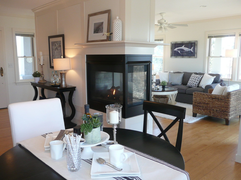 Exemple d'une salle à manger ouverte sur la cuisine bord de mer de taille moyenne avec un mur blanc, parquet clair, une cheminée double-face, un manteau de cheminée en métal et éclairage.