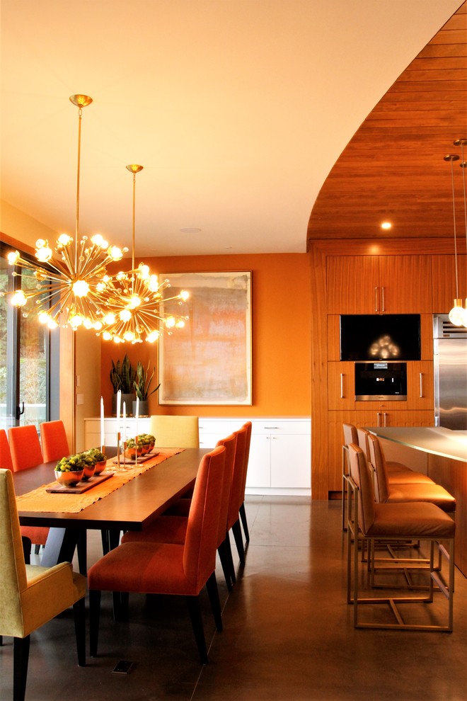 Immagine di un'ampia sala da pranzo aperta verso la cucina moderna con pareti arancioni, pavimento in sughero, camino sospeso, cornice del camino in legno e pavimento grigio