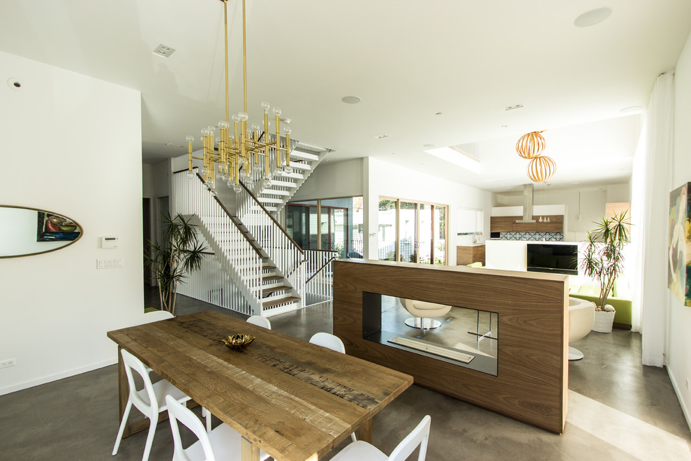 На фото: гостиная-столовая среднего размера в современном стиле с белыми стенами, бетонным полом, двусторонним камином, коричневым полом и фасадом камина из дерева