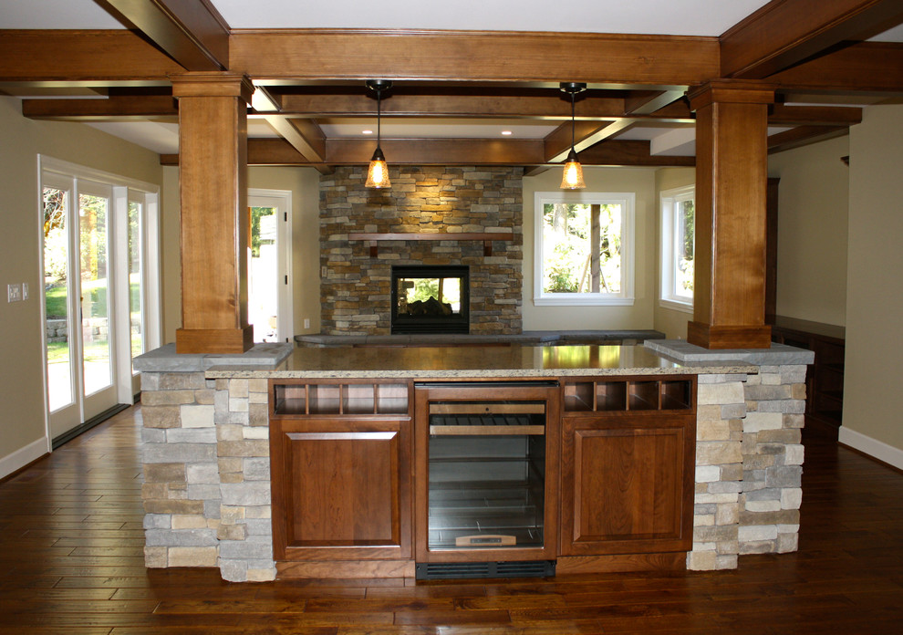 Modelo de comedor de estilo americano grande abierto con paredes beige, suelo de madera oscura, chimenea de doble cara y marco de chimenea de piedra