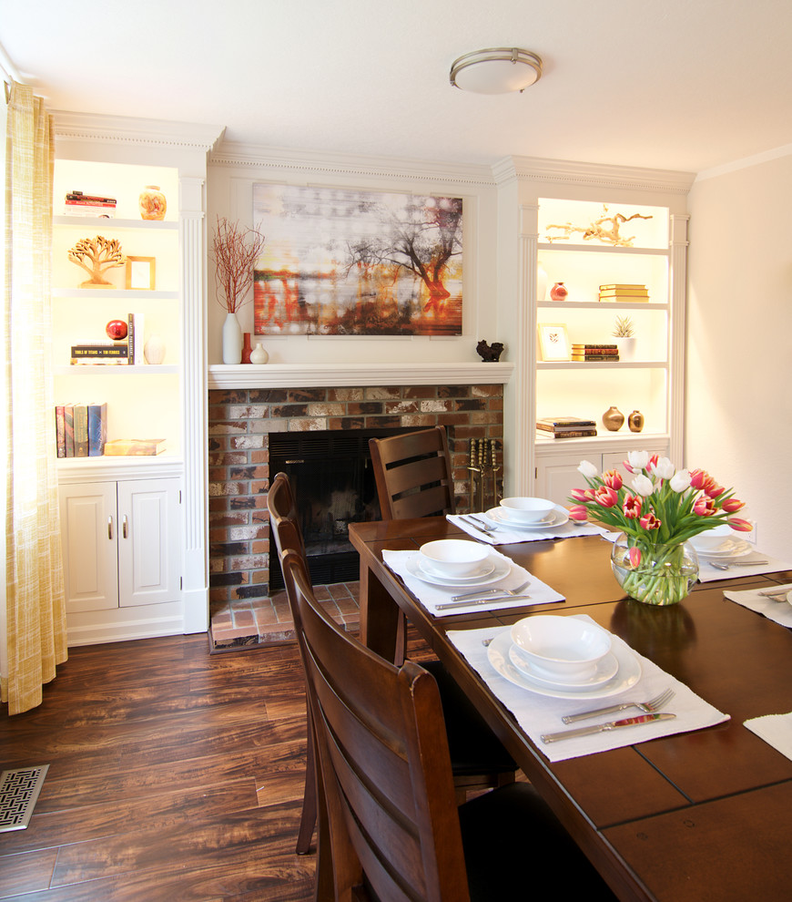 Cette photo montre une salle à manger chic de taille moyenne avec un mur blanc, sol en stratifié, une cheminée standard, un manteau de cheminée en brique et un sol marron.