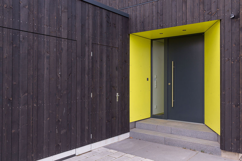 Réalisation d'une porte d'entrée design avec un mur jaune, une porte simple, une porte noire et sol en béton ciré.