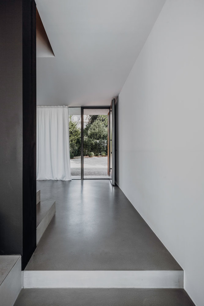 Aménagement d'un très grand hall d'entrée moderne avec un mur blanc, sol en béton ciré, une porte simple, une porte noire et un sol gris.