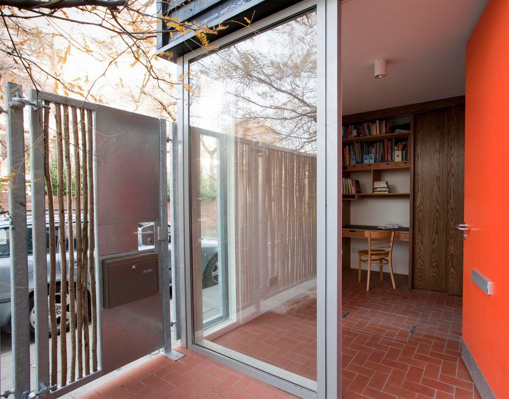 На фото: маленькая узкая прихожая в современном стиле с белыми стенами, кирпичным полом, одностворчатой входной дверью, оранжевой входной дверью и красным полом для на участке и в саду