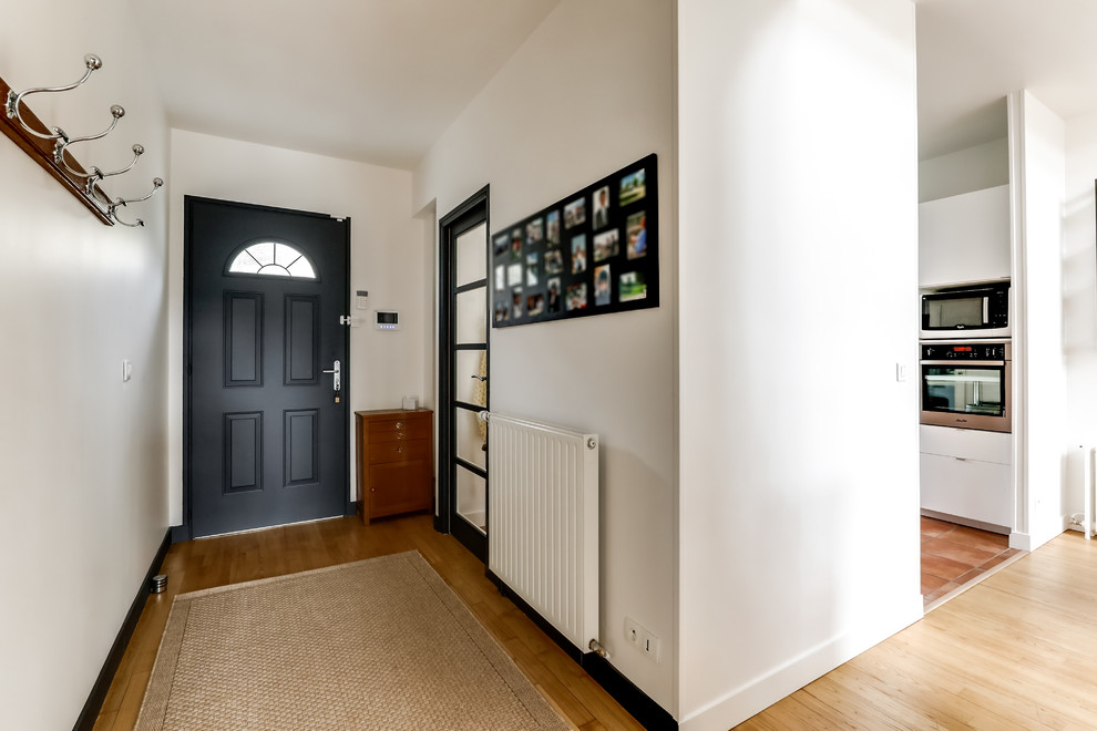 Bild på en mellanstor funkis hall, med vita väggar, mellanmörkt trägolv, en enkeldörr, en svart dörr och beiget golv