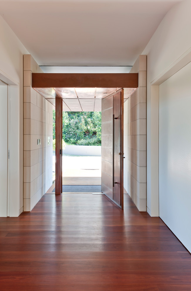 Réalisation d'une entrée minimaliste avec un mur blanc, un sol en bois brun, une porte pivot et une porte en bois brun.