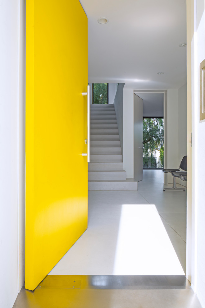 Aménagement d'une entrée avec un mur blanc, une porte pivot, une porte jaune et un sol gris.