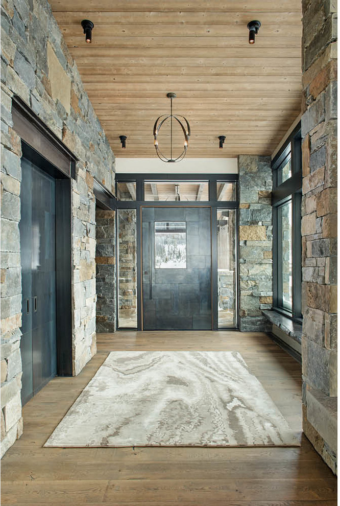 Réalisation d'une grande porte d'entrée chalet avec un mur gris, une porte pivot, une porte métallisée, un sol marron et un sol en bois brun.