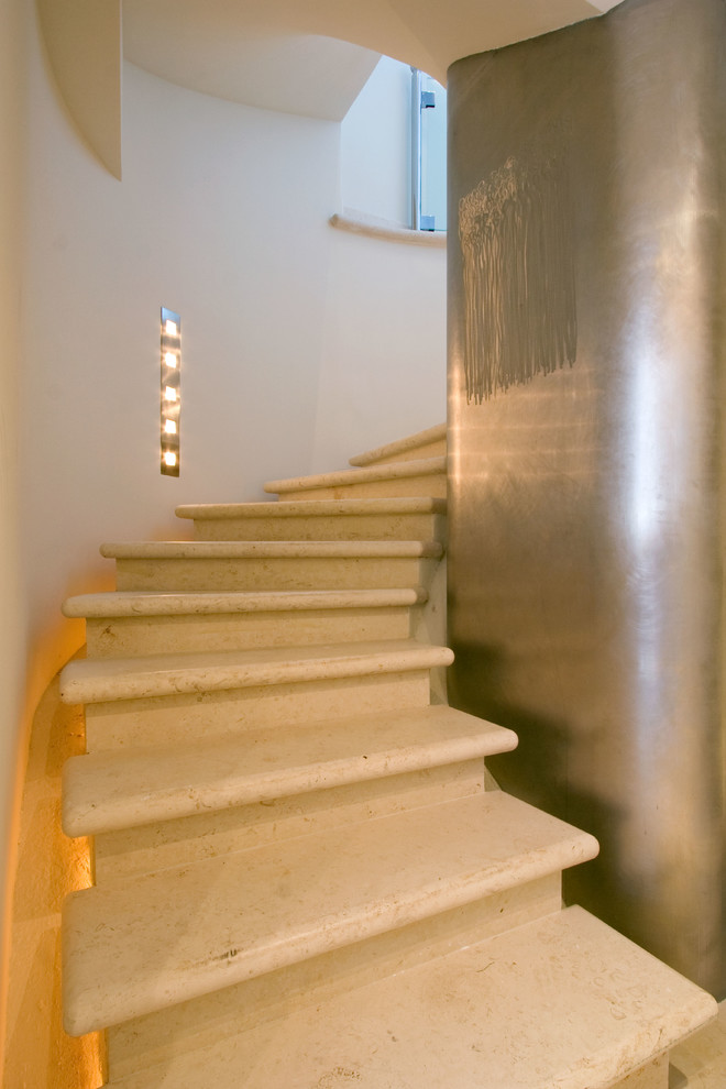Стильный дизайн: большая изогнутая лестница в современном стиле с ступенями из известняка, подступенками из известняка и стеклянными перилами - последний тренд