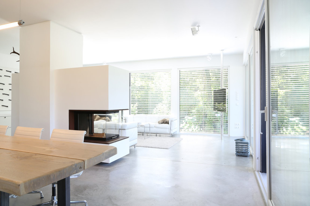Diseño de comedor minimalista abierto con paredes blancas, suelo de cemento, chimenea de doble cara y marco de chimenea de yeso