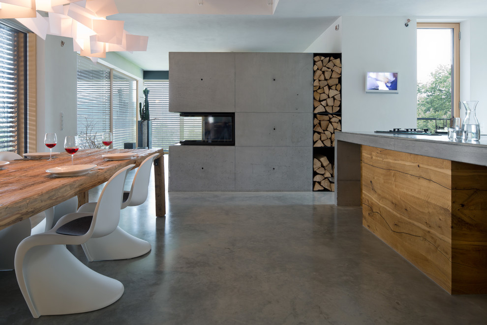 Cette photo montre une salle à manger ouverte sur la cuisine tendance de taille moyenne avec sol en béton ciré, une cheminée double-face, un mur blanc, un manteau de cheminée en béton et un sol gris.