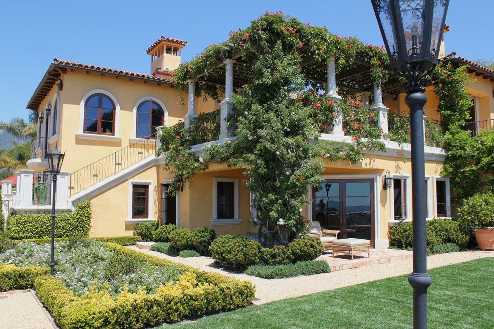 Aménagement d'une grande façade de maison jaune méditerranéenne en stuc à un étage avec un toit à quatre pans.