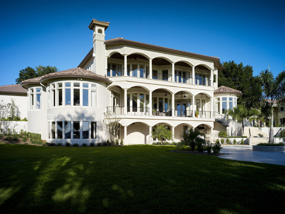 Esempio della villa ampia beige mediterranea a tre piani con rivestimento in stucco, tetto a padiglione e copertura in tegole