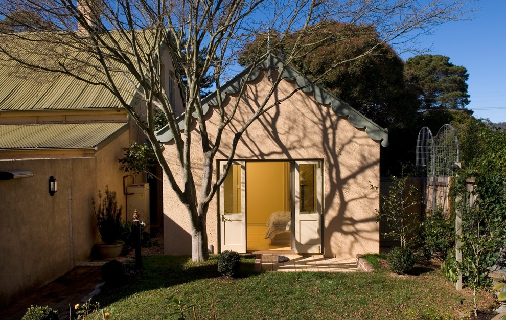 Пример оригинального дизайна: маленький, одноэтажный частный загородный дом в викторианском стиле с облицовкой из цементной штукатурки, двускатной крышей и металлической крышей для на участке и в саду